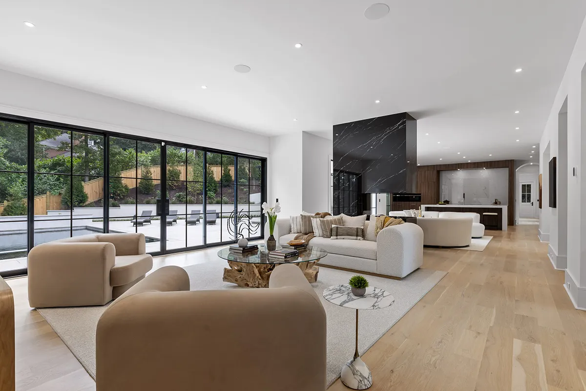 Modern open-concept living room with large floor-to-ceiling sliding glass doors, neutral furniture, light wood flooring, and a view of an outdoor patio with seating and landscaping.